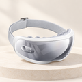 White heated eye massager with a gray front and adjustable strap on a cream display pedestal